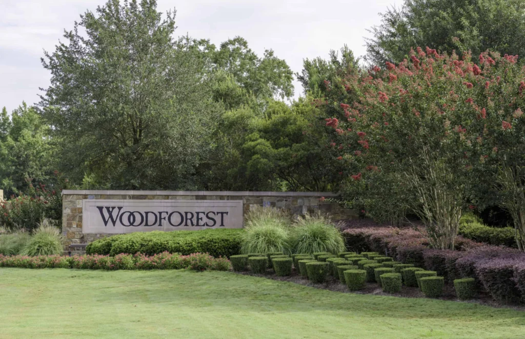 Entrada con letrero de bienvenida a la comunidad Woodforest, rodeada de jardines y vegetación.